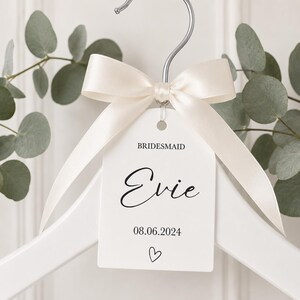 May include: A white wooden clothes hanger featuring a silver metal hook and a cream-colored satin ribbon tied in a bow. A white rectangular tag hangs from the hook, printed with the text BRIDESMAID, the name Evie, the date 08.06.2024, and a heart.