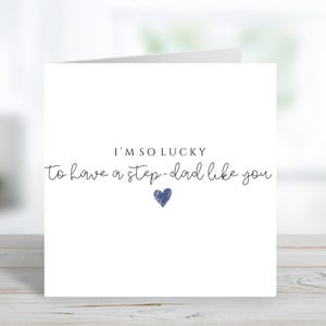 May include: A white card with the message "I'M SO LUCKY to have a step-dad like you" in black script. A small blue heart is beneath the text. The card is on a light wood surface.