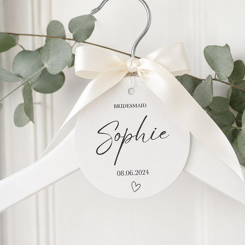 May include: A white wooden clothes hanger featuring a round white tag tied with a cream satin ribbon. The tag displays the text BRIDESMAID, the name Sophie in script, the date 08.06.2024, and a small heart outline printed in black ink.