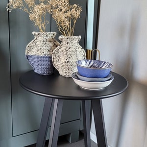 Könnte beinhalten: Zwei gehäkelte Vasen mit getrockneten Blumen auf einem schwarzen Beistelltisch mit drei Beinen. Der Tisch steht neben einem dunkelgrünen Schrank mit einem braunen Ledergriff.