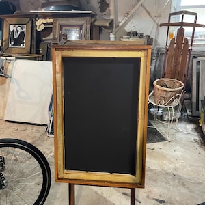 Puede incluir: Una pizarra con marco de madera y superficie negra, sostenida por dos patas. El marco tiene un acabado marrón claro con un borde interior dorado. La pizarra está en una habitación con otros objetos, incluyendo una rueda de bicicleta y cuadros enmarcados.