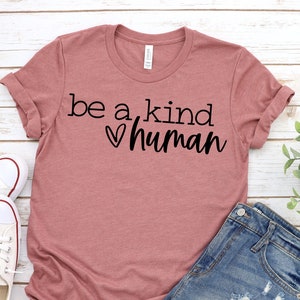 May include: A mauve t-shirt with the text "be a kind human" and a heart symbol. The shirt is laid flat on a white wooden surface with a pair of white sneakers and denim shorts.