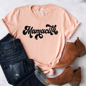 Pu&ograve; includere: Una maglietta color pesca con la scritta "Mamacita" in nero, con un lettering in stile retr&ograve;. La maglietta &egrave; abbinata a jeans in denim scuro e stivaletti marroni. Gli articoli sono disposti su una superficie di marmo bianco.