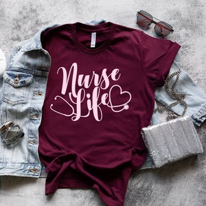 K&ouml;nnte beinhalten: Ein maroonfarbenes T-Shirt mit dem Text "Nurse Life" in rosa Schrift und einer Stethoskopgrafik. Das Shirt liegt flach auf einer grauen, strukturierten Oberfl&auml;che mit einer Jeansjacke, wei&szlig;en Turnschuhen, einer Sonnenbrille und einer silbernen Handtasche.