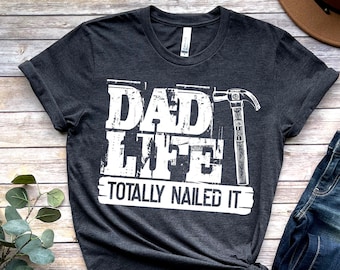 Op de afbeelding: Een donkergrijs T-shirt met een witte afbeelding waarop staat "Dad Life Totally Nailed It" met een hamerafbeelding.