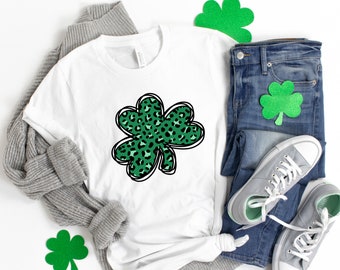May include: A white t-shirt with a green shamrock design featuring a leopard print pattern. The shamrock is outlined in black. The t-shirt is laid out on a white surface with a gray sweater, blue jeans, and green shamrock decorations.