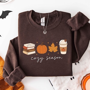 May include: A dark brown long-sleeve sweatshirt featuring a graphic print of a stack of three books, an orange pumpkin, a yellow maple leaf, and a coffee cup with whipped cream. The text "cozy season" is printed in white cursive script below.