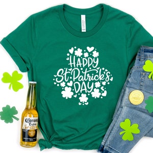 May include: A green t-shirt with the text "Happy St. Patrick's Day" in white, surrounded by shamrocks and hearts. The shirt is paired with blue jeans, shamrock cutouts, and a bottle of beer.