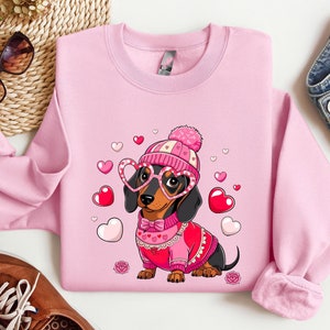 May include: Pink sweatshirt with a cartoon dachshund wearing a pink and white hat and heart-shaped glasses. The dachshund is wearing a red sweater with a white collar and a bow tie. The sweatshirt has a Valentine's Day theme with hearts scattered around the dachshund.