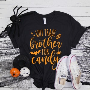 Puede incluir: Camiseta negra con el texto "WILL TRADE brother FOR candy" en naranja. La camiseta est&aacute; combinada con zapatillas blancas, una ara&ntilde;a negra y decoraciones de Halloween. Un dise&ntilde;o divertido y festivo para la temporada de Halloween.
