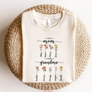 May include: A white t-shirt with a floral design and the text "First Mom Now Grandma" with names listed below each flower.