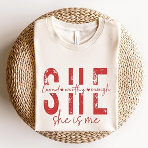 May include: A white t-shirt with a red graphic that says "She is loved, worthy, enough. She is me." The graphic has a distressed look.