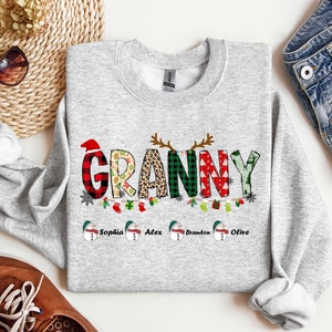 May include: A gray Christmas sweatshirt with the word "Granny" spelled out in colorful patchwork letters. The letters are decorated with snowflakes, Christmas lights, and ornaments. Below the word "Granny" are four snowmen with the names "Sophia", "Alex", "Brandon", and "Olive" written underneath.