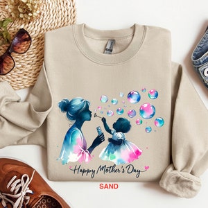 May include: A beige sweatshirt with a watercolor illustration of a mother and child blowing bubbles. The text "Happy Mother's Day" is printed in pink and the word "SAND" is printed in black.