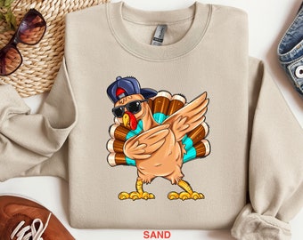 Peut inclure: Un sweat-shirt beige avec une image de dessin animé d'une dinde portant des lunettes de soleil et une casquette de baseball à l'envers. La dinde tient une bière dans chaque aile et a un motif de plumes bleu et turquoise. Le texte "SAND" est imprimé sous l'image.