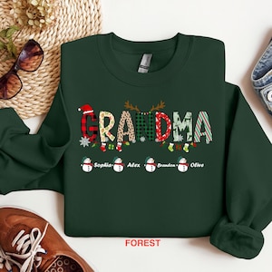 May include: Green Christmas sweatshirt with the word "Grandma" in colorful letters, decorated with snowflakes, reindeer antlers, and snowmen. The names "Sophia", "Alex", "Brandon", and "Olive" are printed below the word "Grandma". The sweatshirt is on a white background.