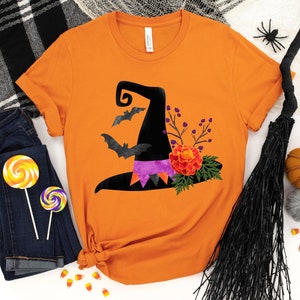 May include: Orange t-shirt with a black witch hat, bats, and floral design. The hat has a purple and black ribbon.