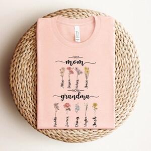 May include: Pink t-shirt with the text "First Mom - Now Grandma" and floral designs. The names Alex, Jessi, Kelsey, Jace, Madeline, James, Daisy, Vaylen, and Jack are written below the flowers.