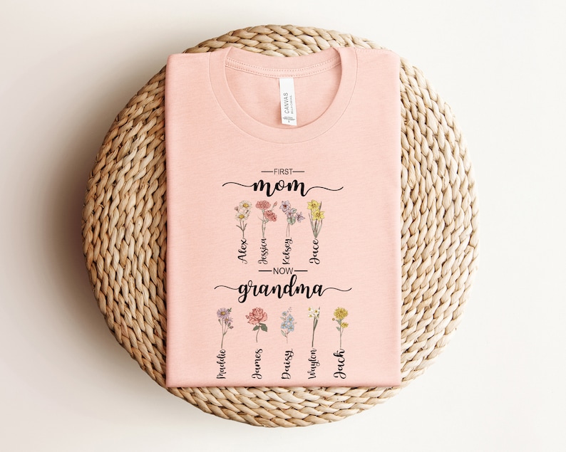 May include: Pink t-shirt with the text "First Mom - Now Grandma" and floral designs. The names Alex, Jessi, Kelsey, Jace, Madeline, James, Daisy, Vaylen, and Jack are written below the flowers.