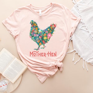 Puede incluir: Una camiseta rosa claro con un dise&ntilde;o floral de gallina. La gallina est&aacute; compuesta por flores coloridas. El texto "MOTHER HEN" est&aacute; impreso en coral debajo. La camiseta est&aacute; combinada con un libro, un melocot&oacute;n y una pajita.