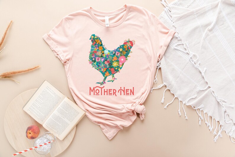 Puede incluir: Una camiseta rosa claro con un dise&ntilde;o floral de gallina. La gallina est&aacute; compuesta por flores coloridas. El texto "MOTHER HEN" est&aacute; impreso en coral debajo. La camiseta est&aacute; combinada con un libro, un melocot&oacute;n y una pajita.