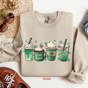 May include: A light beige sweatshirt with a graphic of four green shamrock-themed drinks. The drinks are labeled "Lucky a Latte" and have whipped cream and straws. The sweatshirt is labeled "SAND".