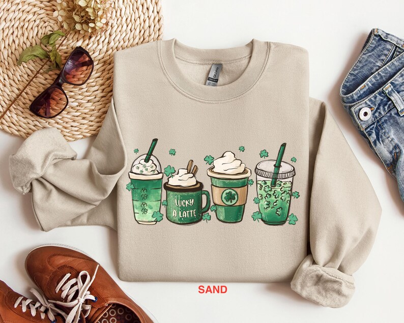 May include: A light beige sweatshirt with a graphic of four green shamrock-themed drinks. The drinks are labeled "Lucky a Latte" and have whipped cream and straws. The sweatshirt is labeled "SAND".
