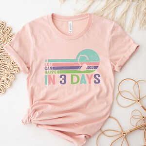 Puede incluir: Una camiseta rosa con un gr&aacute;fico de estilo retro que dice "A lot can happen in 3 days". El gr&aacute;fico presenta un dise&ntilde;o a rayas de colores con un c&iacute;rculo en el centro.