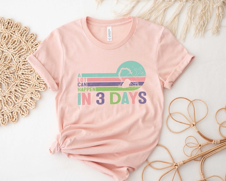 Puede incluir: Una camiseta rosa con un gr&aacute;fico de estilo retro que dice "A lot can happen in 3 days". El gr&aacute;fico presenta un dise&ntilde;o a rayas de colores con un c&iacute;rculo en el centro.