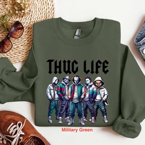 Peut inclure: Sweat-shirt vert militaire avec le texte "THUG LIFE" en noir. Le graphisme pr&eacute;sente cinq personnages en sweats &agrave; capuche et vestes. Le sweat-shirt est associ&eacute; &agrave; des chaussures marron et un jean bleu.