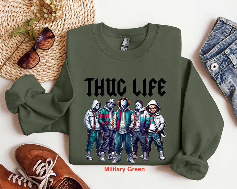 Peut inclure: Sweat-shirt vert militaire avec le texte "THUG LIFE" en noir. Le graphisme pr&eacute;sente cinq personnages en sweats &agrave; capuche et vestes. Le sweat-shirt est associ&eacute; &agrave; des chaussures marron et un jean bleu.
