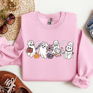 May include: Pink sweatshirt with a graphic of five cartoon ghosts dressed as nurses. The ghosts are holding medical supplies like a syringe, a first aid kit, and a clipboard. The text "Boo Crew" is printed on the sweatshirt.