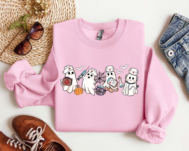 May include: Pink sweatshirt with a graphic of five cartoon ghosts dressed as nurses. The ghosts are holding medical supplies like a syringe, a first aid kit, and a clipboard. The text "Boo Crew" is printed on the sweatshirt.