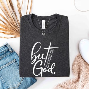 May include: Dark gray t-shirt with the text "but God" in white script and a cross graphic. The shirt also has the text "There was no way, but GOD made a way." A silver necklace with a sun pendant is on the shirt.