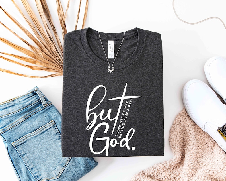 May include: Dark gray t-shirt with the text "but God" in white script and a cross graphic. The shirt also has the text "There was no way, but GOD made a way." A silver necklace with a sun pendant is on the shirt.
