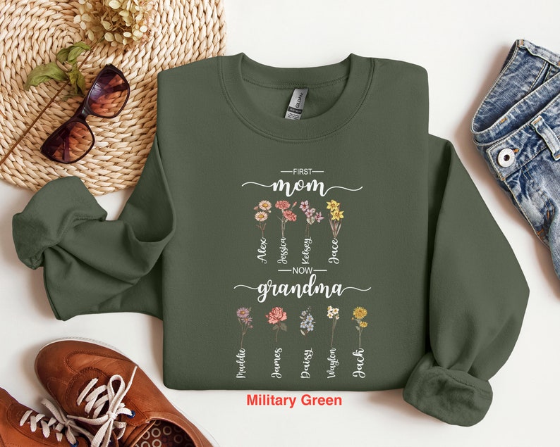 May include: A military green sweatshirt with the text "First Mom Now Grandma" and floral designs. The text is surrounded by names, including Alex, Jessica, Kelsey, Grace,  and more.