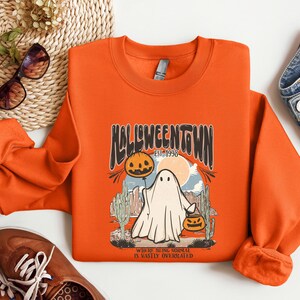 May include: Orange sweatshirt with a retro Halloween design featuring a ghost holding a jack-o-lantern. The text reads "Halloweentown Est. 1998" and "Where being normal is vastly overrated".
