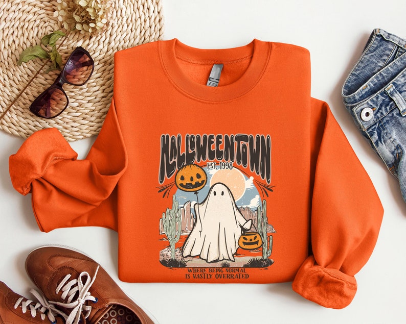 May include: Orange sweatshirt with a retro Halloween design featuring a ghost holding a jack-o-lantern. The text reads "Halloweentown Est. 1998" and "Where being normal is vastly overrated".