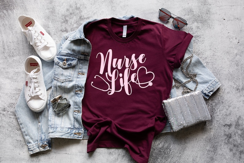 K&ouml;nnte beinhalten: Ein maroonfarbenes T-Shirt mit dem Text "Nurse Life" in rosa Schrift und einer Stethoskopgrafik. Das Shirt liegt flach auf einer grauen, strukturierten Oberfl&auml;che mit einer Jeansjacke, wei&szlig;en Turnschuhen, einer Sonnenbrille und einer silbernen Handtasche.