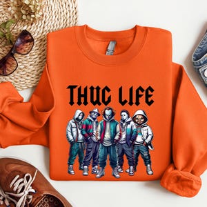 Peut inclure: Sweat-shirt orange avec l'inscription "THUG LIFE" en noir. Le sweat-shirt pr&eacute;sente un graphisme de cinq personnages de dessins anim&eacute;s portant des sweats &agrave; capuche et des vestes. Les personnages sont dans des tons de bleu, vert et blanc.