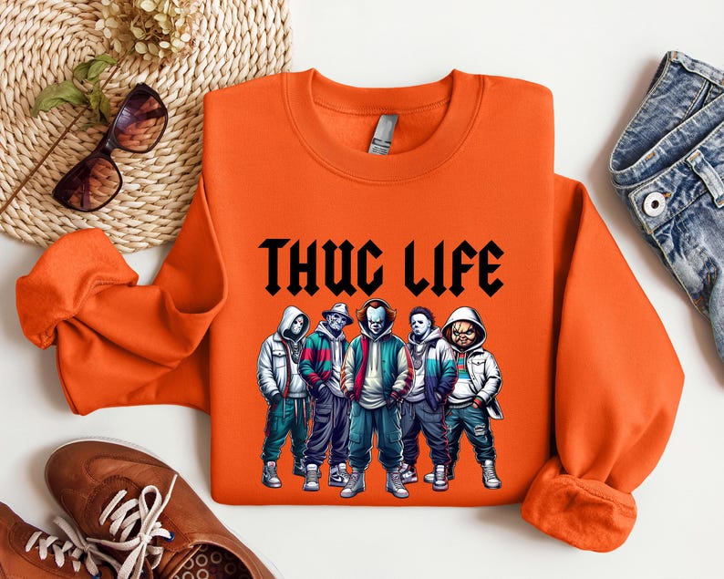 Peut inclure: Sweat-shirt orange avec l'inscription "THUG LIFE" en noir. Le sweat-shirt pr&eacute;sente un graphisme de cinq personnages de dessins anim&eacute;s portant des sweats &agrave; capuche et des vestes. Les personnages sont dans des tons de bleu, vert et blanc.