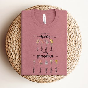 May include: A pink t-shirt with a floral design and the text "First Mom - Now Grandma" with names listed below.
