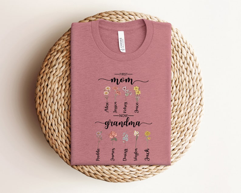 May include: A pink t-shirt with a floral design and the text "First Mom - Now Grandma" with names listed below.