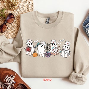 May include: A tan sweatshirt with a graphic of five ghosts dressed as nurses. The ghosts are holding medical supplies like a syringe, a first aid kit, and a clipboard. The text "SAND" is printed on the bottom of the sweatshirt.