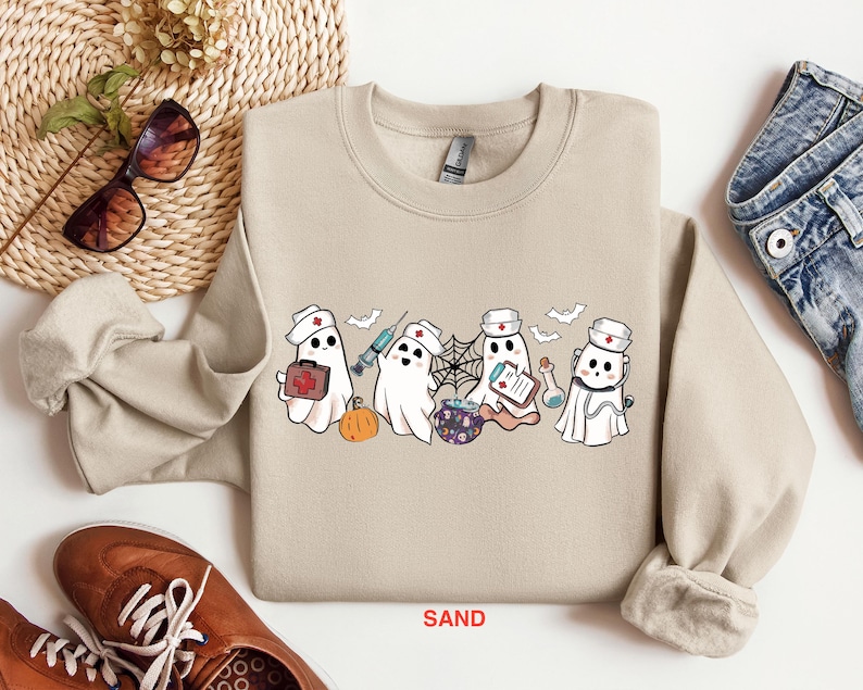 May include: A tan sweatshirt with a graphic of five ghosts dressed as nurses. The ghosts are holding medical supplies like a syringe, a first aid kit, and a clipboard. The text "SAND" is printed on the bottom of the sweatshirt.