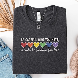 May include: Dark gray t-shirt with the text "BE CAREFUL WHO YOU HATE, it could be someone you love." The text is above a row of six rainbow-colored hearts. A silver necklace with a sun pendant is on the shirt.