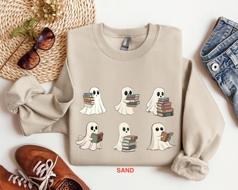 Może przedstawiać: Beżowa bluza z nadrukiem sześciu duch&oacute;w trzymających stosy książek. Na dole bluzy znajduje się napis "SAND".
