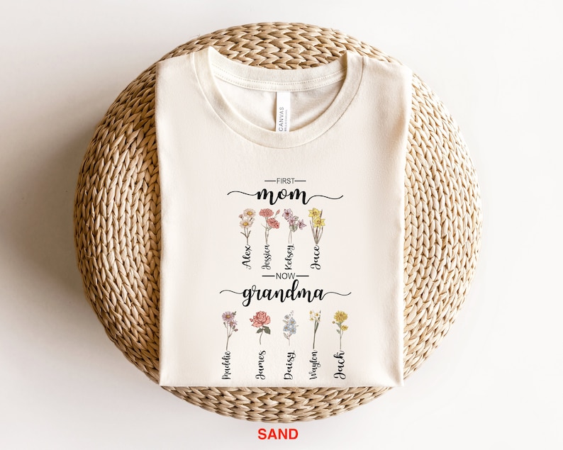 May include: A white t-shirt with a floral design and the text "First Mom Now Grandma" with names listed below each flower.