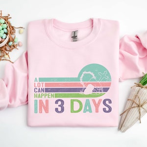 Puede incluir: Sudadera rosa con un gr&aacute;fico de estilo retro que dice "A LOT CAN HAPPEN IN 3 DAYS". El gr&aacute;fico presenta una imagen estilizada de una tumba con una cruz y una salida del sol.