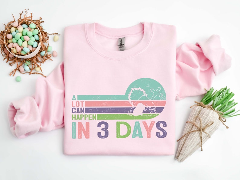 Puede incluir: Sudadera rosa con un gr&aacute;fico de estilo retro que dice "A LOT CAN HAPPEN IN 3 DAYS". El gr&aacute;fico presenta una imagen estilizada de una tumba con una cruz y una salida del sol.